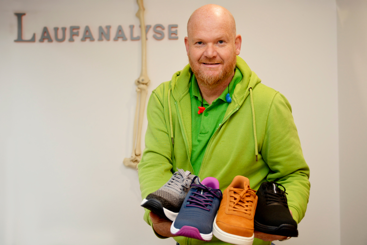 Mann mit grüner Jacke hält vier verschiedene Schuhmodelle in der Hand